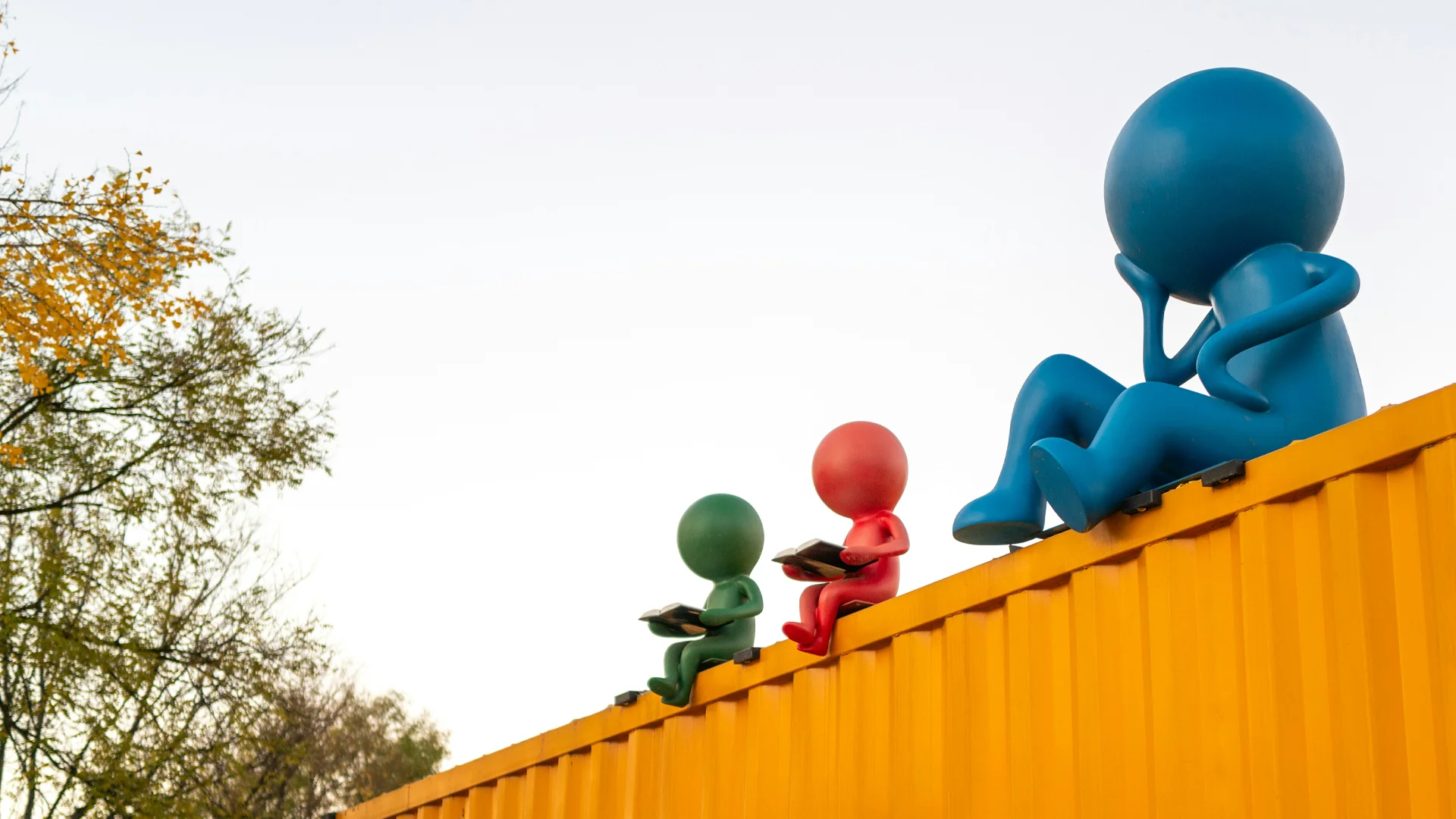 Ein gelber Container in der Natur, auf dem drei bunte Plastikfiguren sitzen, von denen eine nachdenkt und die anderen beiden lesen.